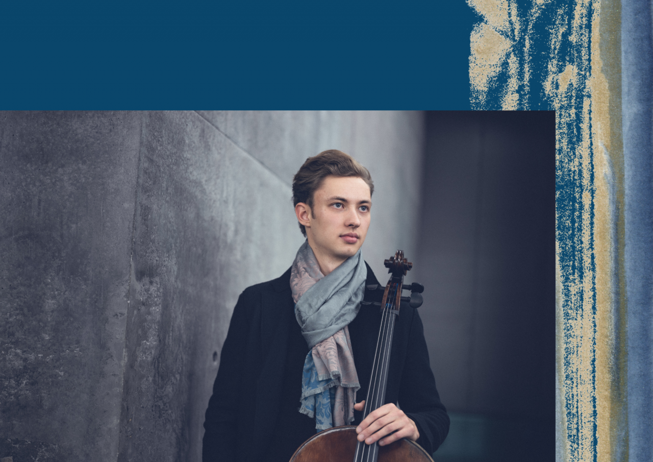 Ein Mann mit Halstuch steht vor einer Betonwand und hält ein Cello.