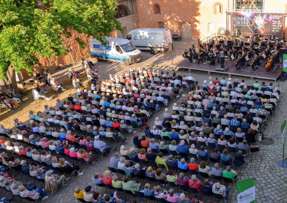 In einem gepflasterten Hof sitzen Zuschauer vor einer kleinen Bühne, auf der ein Orchester spielt.
